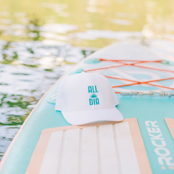 White cap with 'All Day' logo on a surfboard with blurred greenery in the background