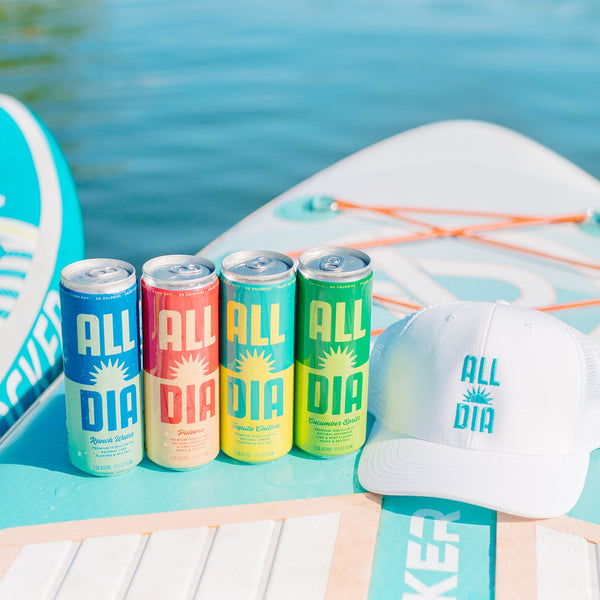 Four cans of All Dia drinks and a cap on a beach setting with water in the background.